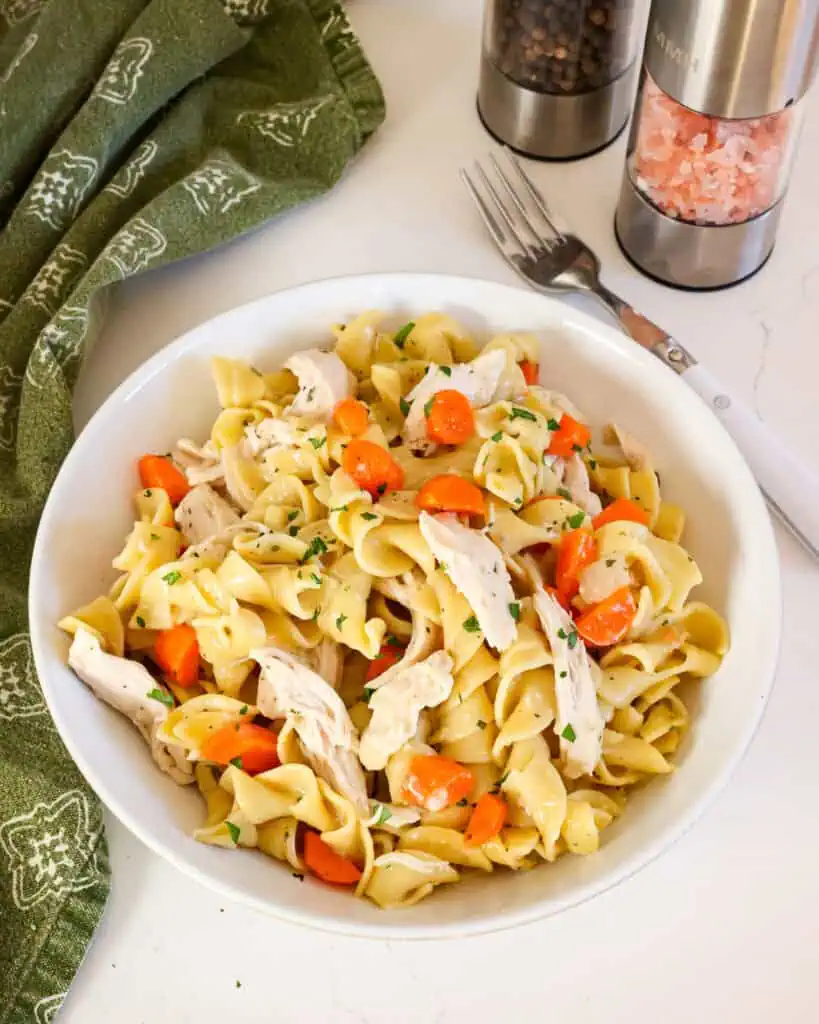 An individual serving bowl full of chicken and noodles with a fork, salt shaker, and pepper shaker in the background. 