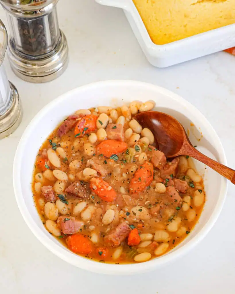 A single serving bowl of ham an beans with a salt and pepper shaker and cornbread in the background. 