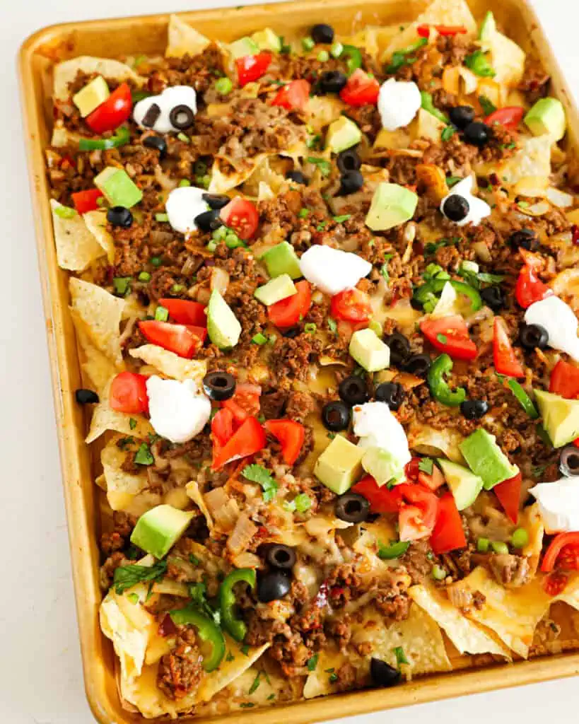 A large baking sheet full of nacho chips layered with seasoned ground beef, queso quesadilla, black olives, tomatoes, jalapeno rings, and sour cream. 
