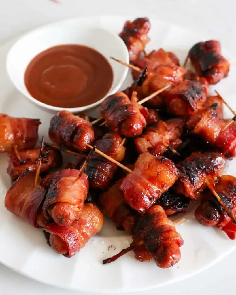 A white plate full of bacon wrapped little smokies basted with brown sugar next to a bowl of BBQ sauce. 