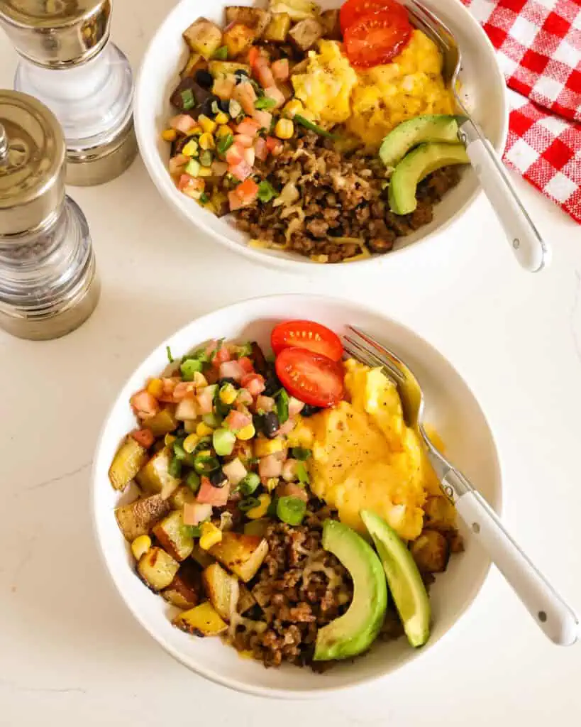 An overhead view of breakfast bowls with fried potatoes, browned sausage, scrambled eggs, and cheese. 