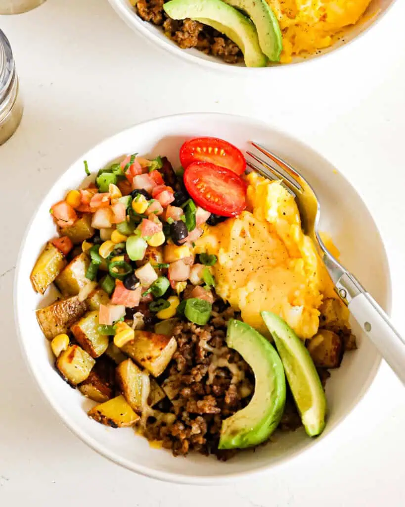 A close up view of a breakfast bowl full of browned pork sausage, fried potatoes. scrambled eggs, Pico de Gallo, and avocado wedges. 