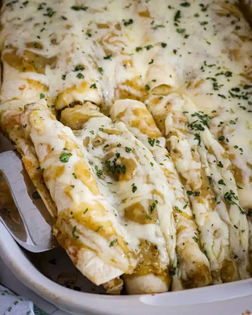 Two green chile chicken enchiladas on a spatula over a dish full green chile chicken enchiladas. 