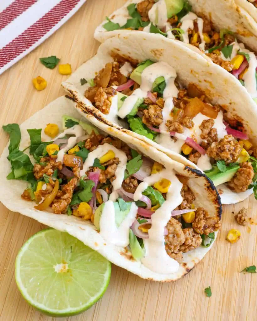 A close up view of two ground pork tacos with blackened corn, lettuce, cilantro, avocado, pickled red onions, and chipotle ranch. 