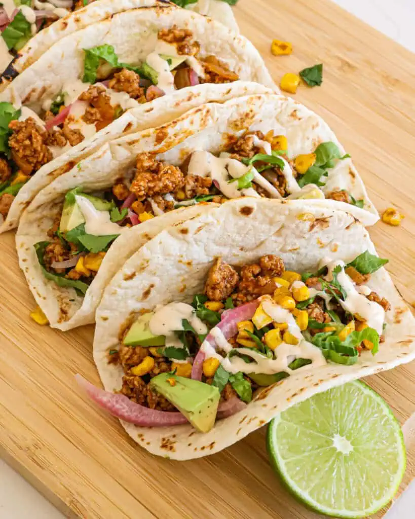 Four ground pork tacos with pickled onions and avocado on a wooden board with fresh limes. 