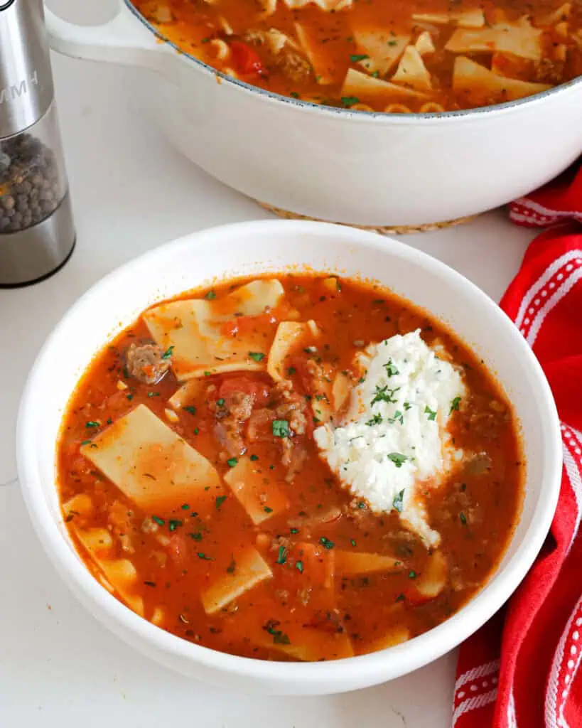 A single serving bowl full of lasagna soup topped with ricotta cheese mixture and chopped fresh parsley in front of a Dutch oven full of lasagna soup. 