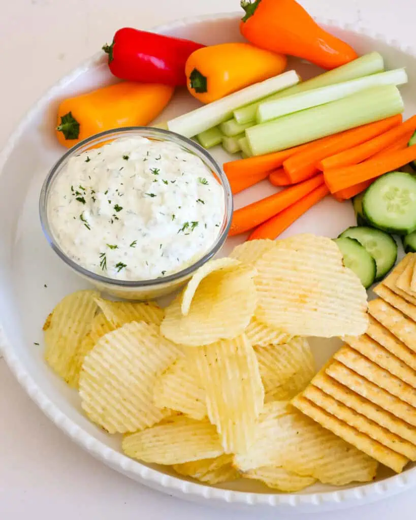 A white round serving platter with a small glass bowl full of dill pickle dip, potato chips, crackers, baby bell peppers, carrot and celery sticks and cucumber slices. 