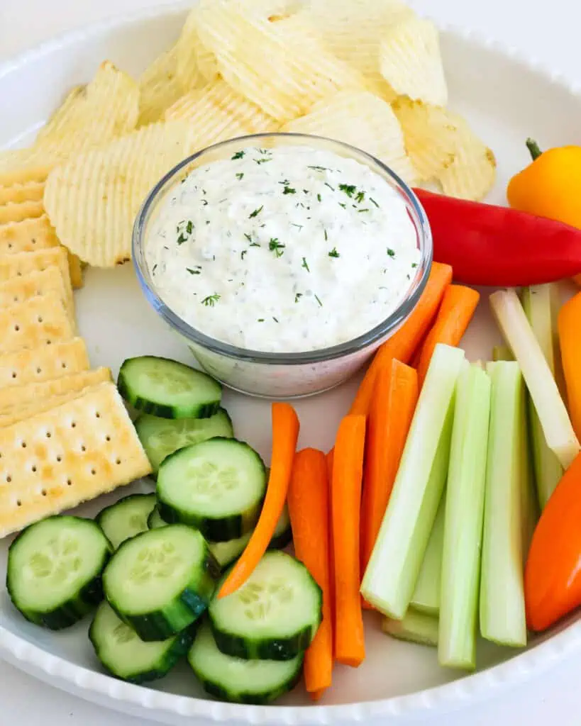 A white round serving platter with a small glass bowl full of dill pickle dip, potato chips, crackers, and assorted vegetables. 