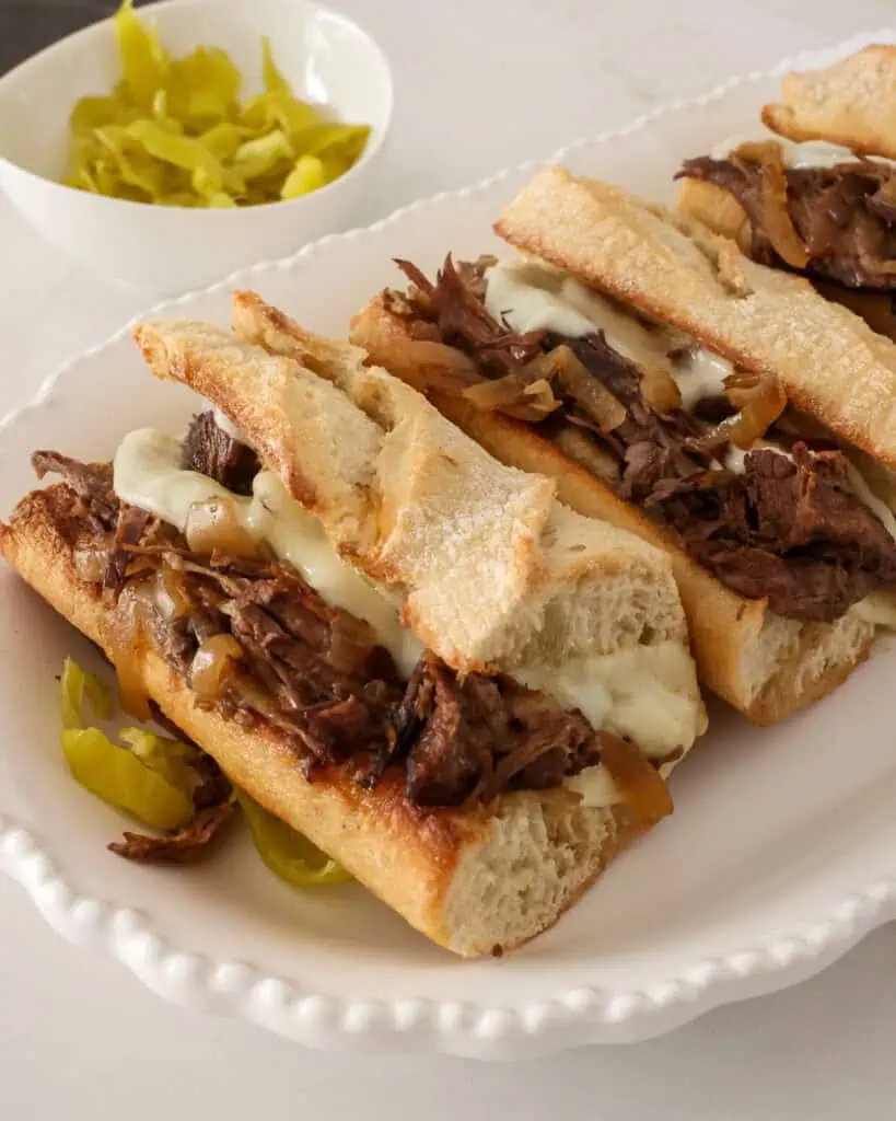 The side view ot two French dip sandwiches on a platter with sliced peppercini peppers. 