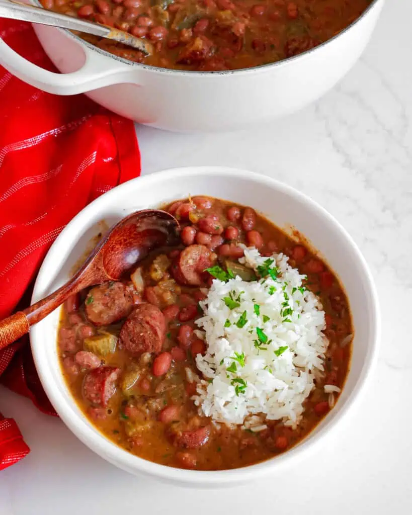 Red beans and rice in a serving bowl with a wooden spoon in front of a Dutch oven filled with red beans and rice. 