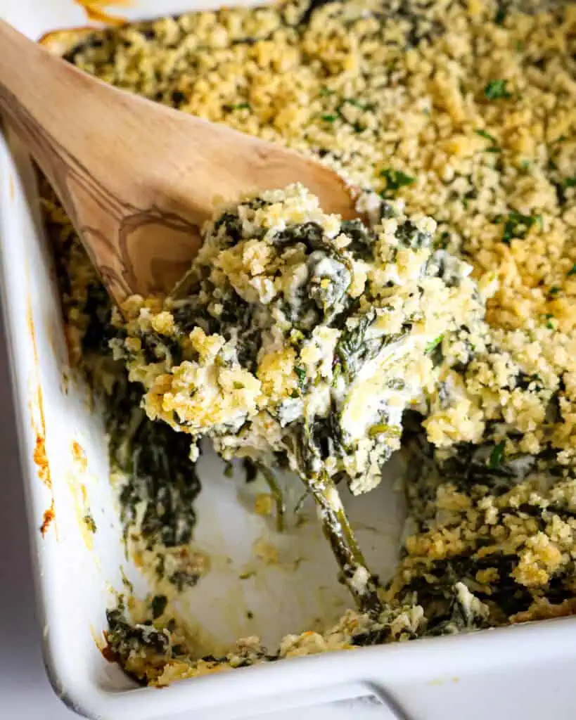 An 8x8-inch white casserole dish and a wooden spoon filled with spinach casserole. 