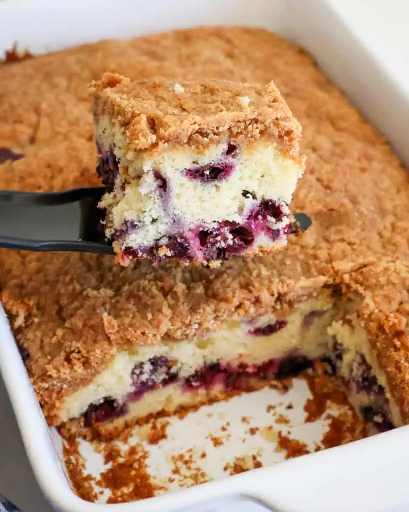 A single piece of blueberry coffee cake on a spatula over the baking dish with the rest of the cake. 