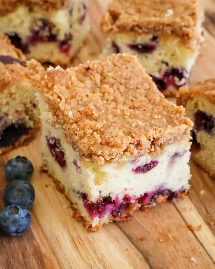 A slice of blueberry coffee cake on a cutting board with a few blueberries on the side. 