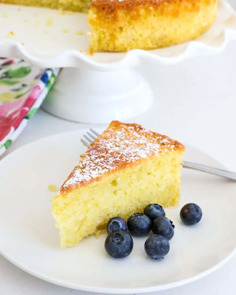 A single serving white plate with a single slice of lemon olive oil cake with a few blueberries on the side. 